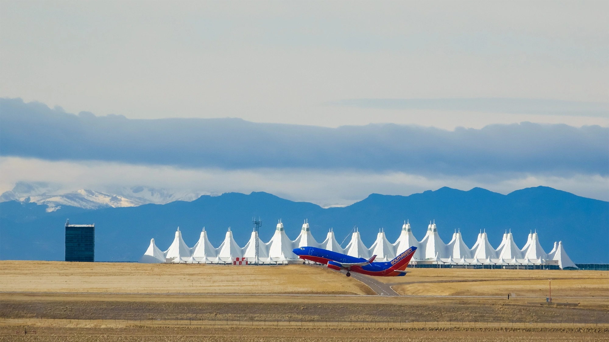 A blue and red aeroplane just taking off from the runway against a backdrop of the airport and snowcapped mountains.