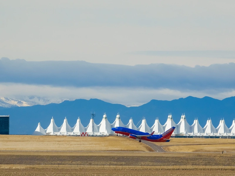 A blue and red aeroplane just taking off from the runway against a backdrop of the airport and snowcapped mountains.