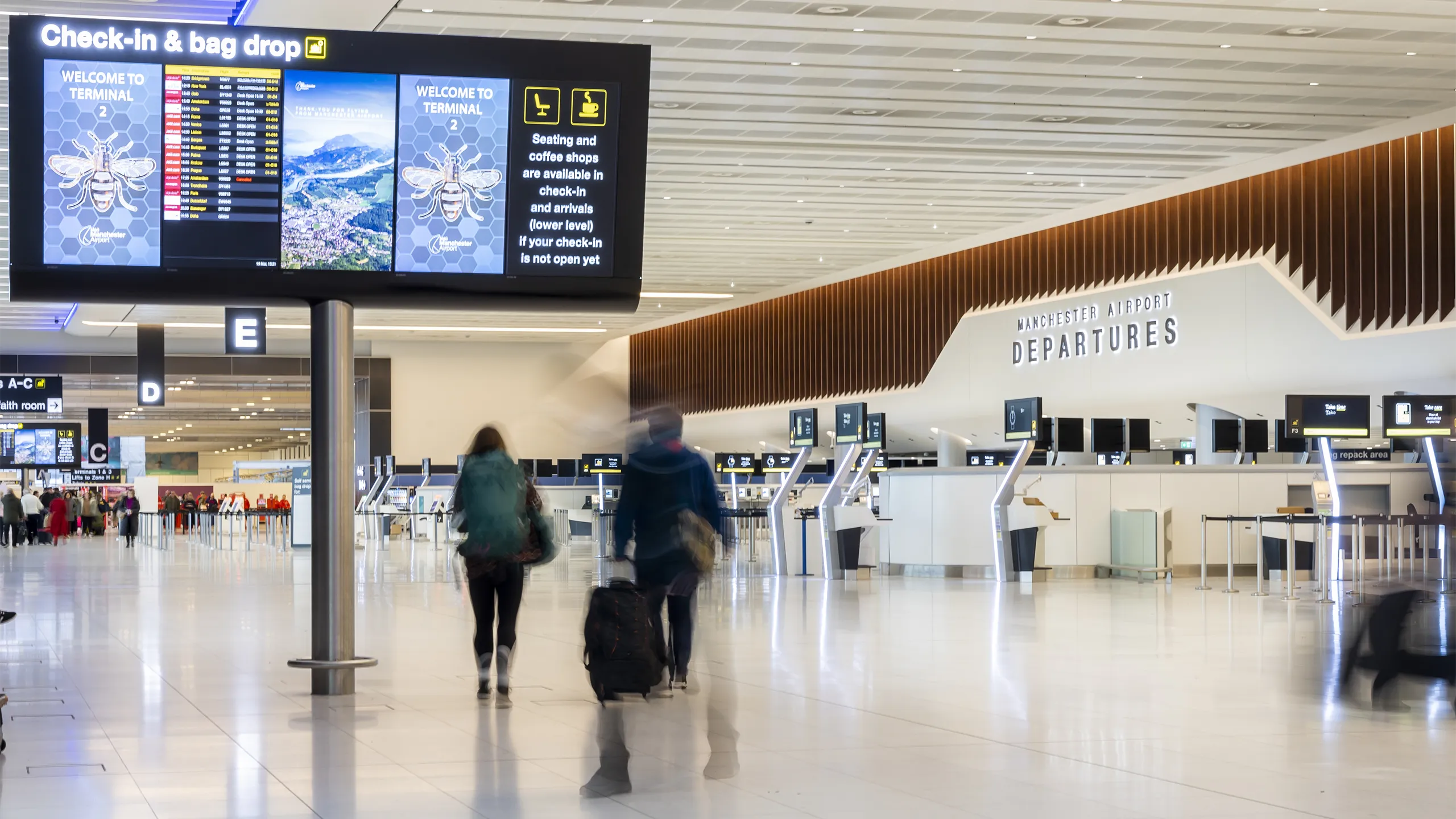 Wide view of an airport departure hall with check in kiosks, digital screens overhead and two travelers walking with luggage.