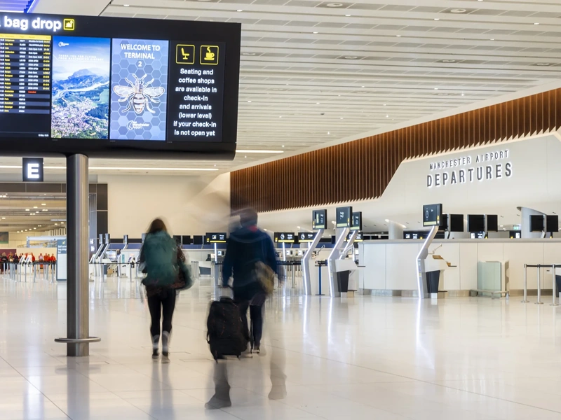 Wide view of an airport departure hall with check in kiosks, digital screens overhead and two travelers walking with luggage.