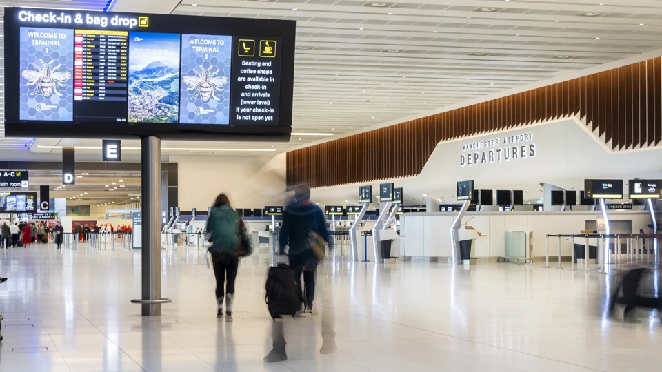 Wide view of an airport departure hall with check in kiosks, digital screens overhead and two travelers walking with luggage.