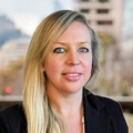 portrait of Caitlin Adair, Director, with long blonde hair, standing outside featuring an urban cityscape with tall buildings and a mix of architectural styles. The scene includes some trees with autumn foliage, adding a touch of color.