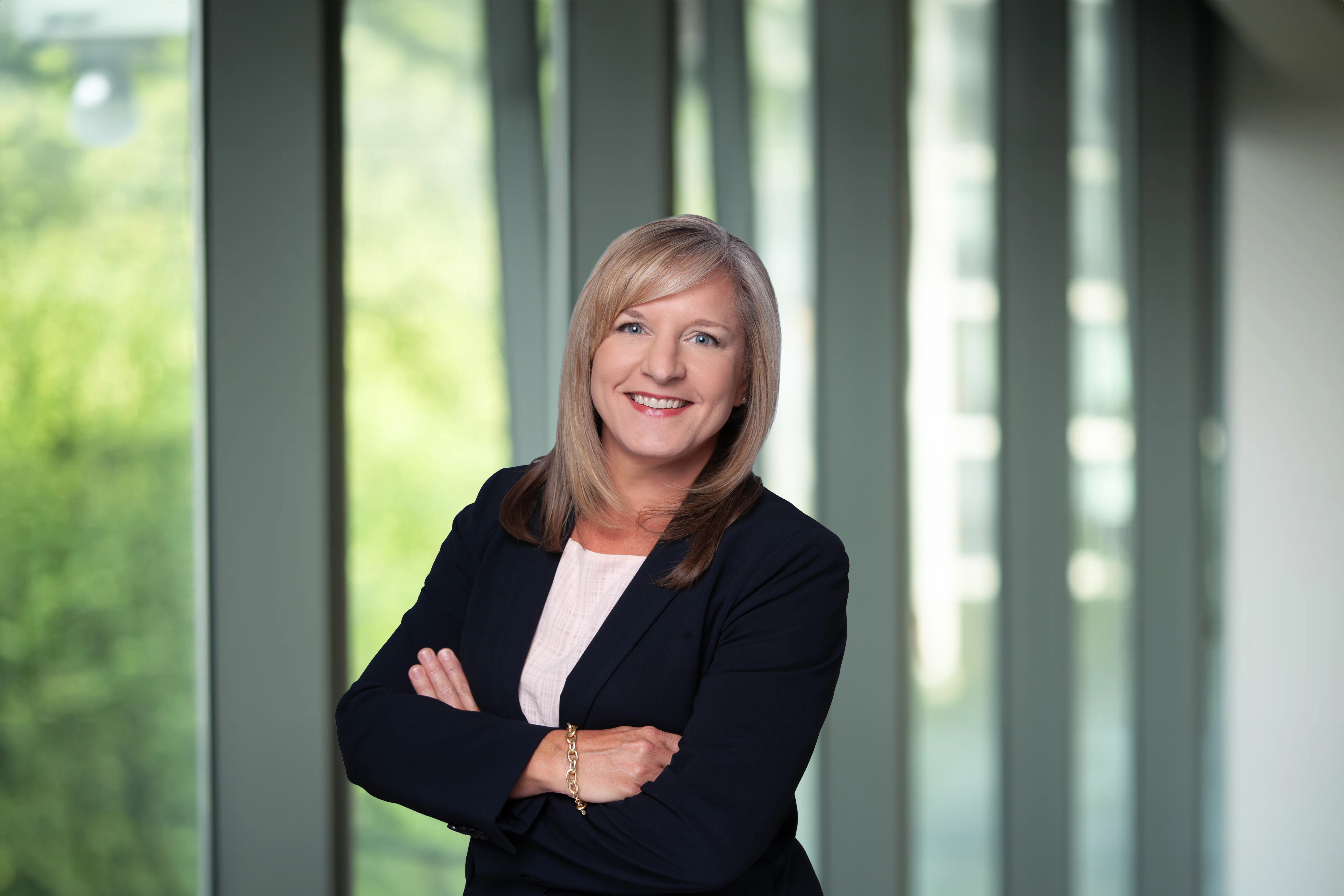Heather McKeen, standing in office in front a window of an outdoor urban setting, featuring modern architectural elements with clean lines. There are hints of greenery, suggesting some trees or plants.