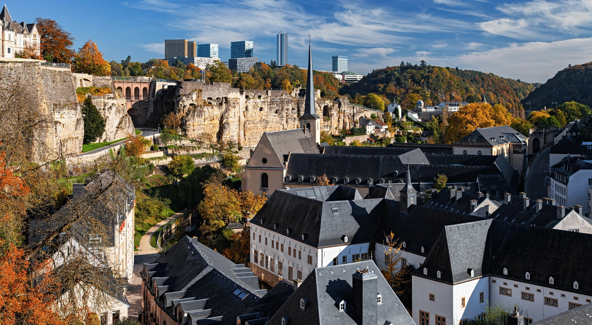 Historic town with stone walls, a church spire and rooftops surrounded by autumn trees.
