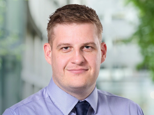 Steven Jackson, UK Director of Infrastructure, standing outside in front of an outdoor urban setting, featuring modern architectural elements with clean lines. There are hints of greenery, suggesting some trees or plants.