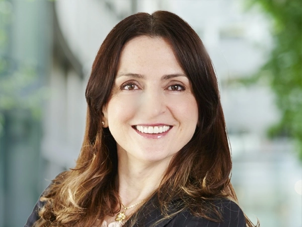 Portrait of Sacira Coric, Clean Energy & Decarbonisation, Global Programme Advisory Lead, standing outside in front of an outdoor urban setting, featuring modern architectural elements with clean lines. There are hints of greenery, suggesting some trees or plants.