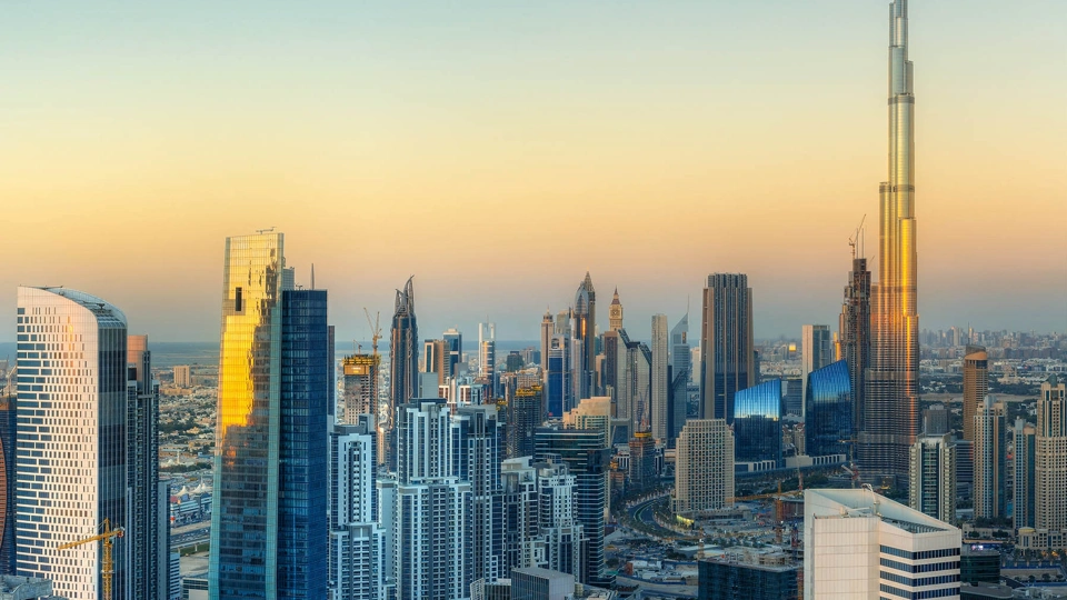 A panoramic view of a modern city skyline at sunset, featuring numerous high-rise buildings and skyscrapers, including the iconic Burj Khalifa on the right, dominating the landscape with its towering height. The buildings are illuminated by the warm golden light of the setting sun, creating a striking contrast against the clear sky.