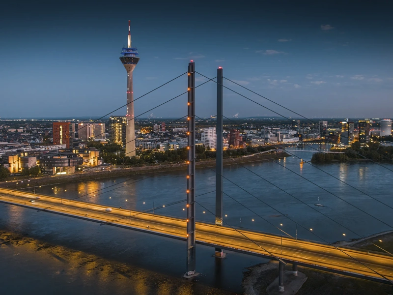 Aerial blue-hour view of the Düsseldorf skyline along the Rhine River, featuring the illuminated Rheinkniebrücke leading toward the city center and the Rheinturm rising above modern buildings. City lights reflect on the calm water, creating a clean, contemporary European cityscape. North Rhine–Westphalia, Germany, Europe.