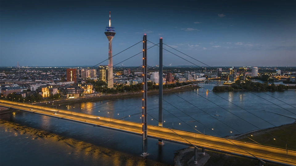 Aerial blue-hour view of the Düsseldorf skyline along the Rhine River, featuring the illuminated Rheinkniebrücke leading toward the city center and the Rheinturm rising above modern buildings. City lights reflect on the calm water, creating a clean, contemporary European cityscape. North Rhine–Westphalia, Germany, Europe.