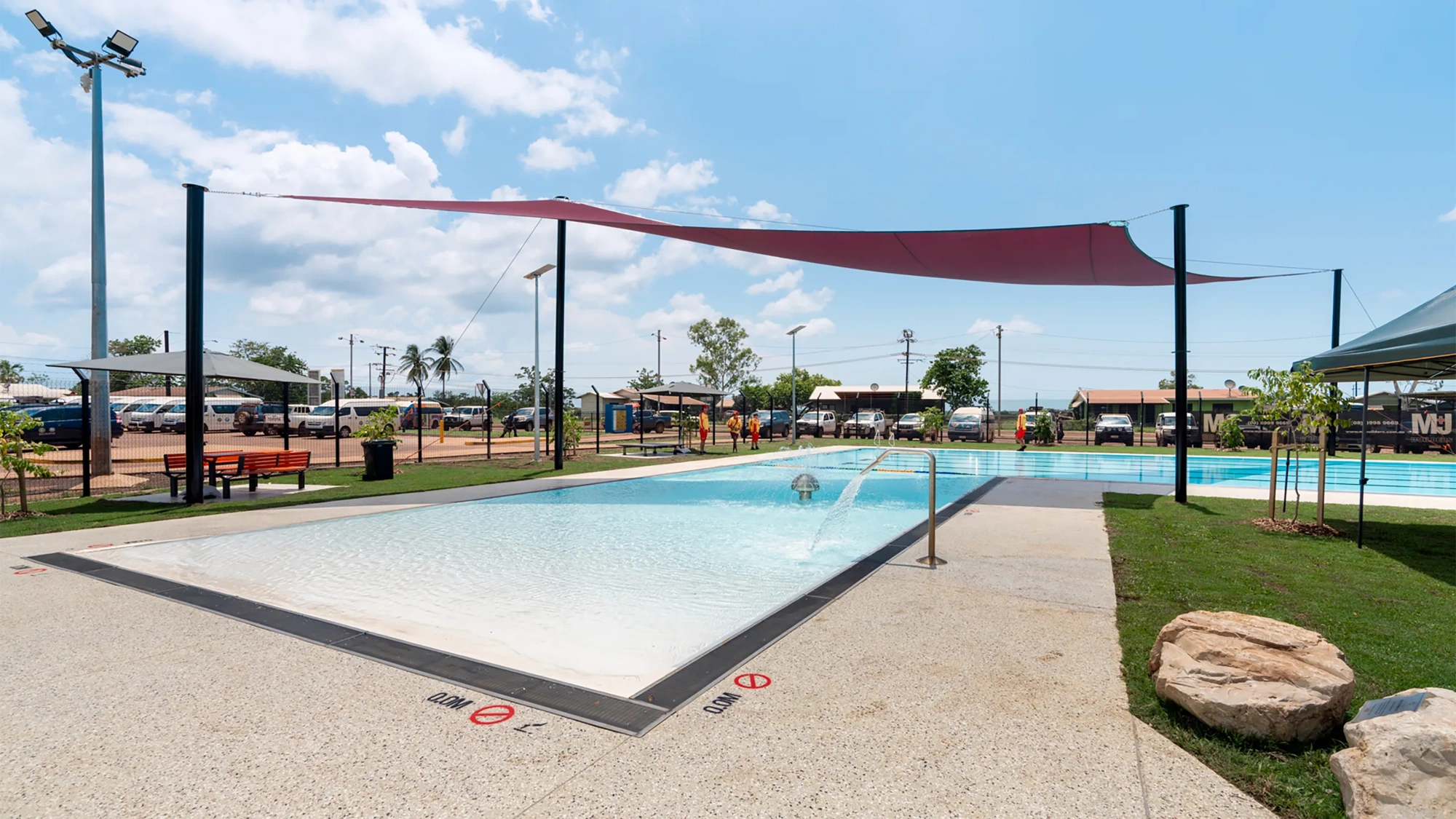 An outdoor swimming pool with shallow water, surrounded by a concrete deck. The pool has a sloped entry and a handrail on the right side. There are signs on the pool deck indicating "No Diving" and depth markings in both feet and meters. Above the pool, a large red sunshade is stretched between several poles to provide shade. The surrounding area includes some green grass, benches, and small trees. In the background, there are buildings, palm trees, and parked cars under a partly cloudy sky.