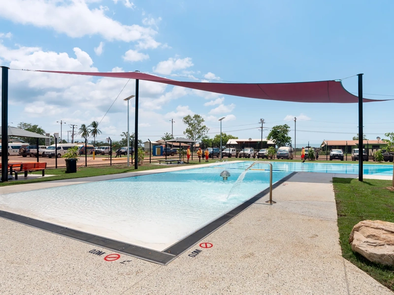 An outdoor swimming pool with shallow water, surrounded by a concrete deck. The pool has a sloped entry and a handrail on the right side. There are signs on the pool deck indicating "No Diving" and depth markings in both feet and meters. Above the pool, a large red sunshade is stretched between several poles to provide shade. The surrounding area includes some green grass, benches, and small trees. In the background, there are buildings, palm trees, and parked cars under a partly cloudy sky.