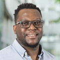 Gerald Madondo, Country Director, standing outside in front of an outdoor urban setting, featuring modern architectural elements with clean lines. There are hints of greenery, suggesting some trees or plants.