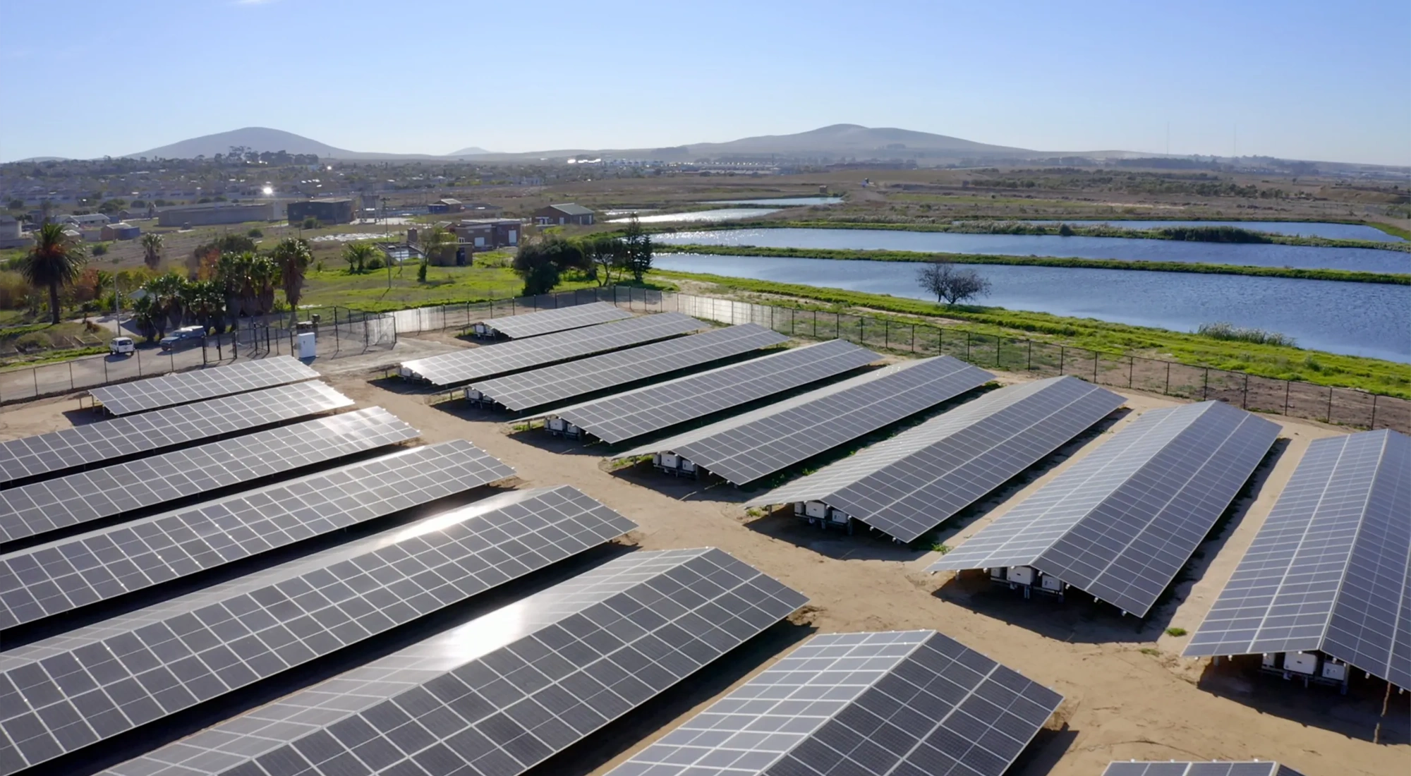 A collection of solar panels next to a body of water with mountains in the background.