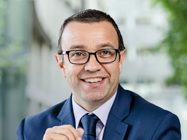 Andy Aston, Global Head of Client Solutions for Natural Resources, standing outside in front of an outdoor urban setting, featuring modern architectural elements with clean lines. There are hints of greenery, suggesting some trees or plants.