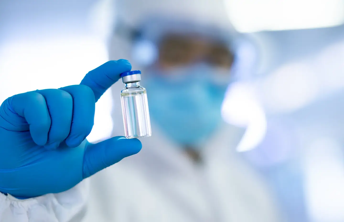 A person dressed in protective laboratory clothing, including blue latex gloves and holding vaccination vile in between their fingers. 