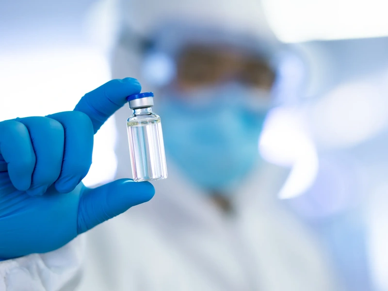 A person dressed in protective laboratory clothing, including blue latex gloves and holding vaccination vile in between their fingers.