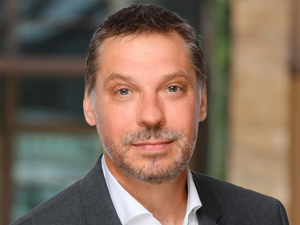 Kirk Smith, Director, Cost and Commercial Management, Germany and Austria,  outside in front of a blurred brown background featuring a building and mix of natural and urban elements.