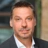 Kirk Smith, Director, Cost and Commercial Management, Germany and Austria, outside in front of a blurred brown background featuring a building and mix of natural and urban elements.