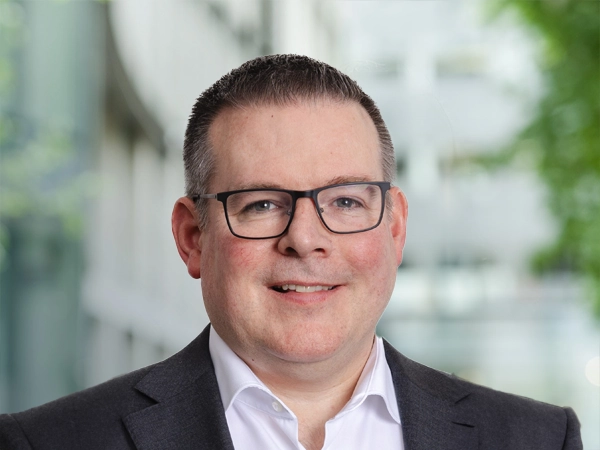  Portrait of Euan Reaper, Director, Europe, Cost Management, standing outside in front of an outdoor urban setting, featuring modern architectural elements with clean lines. There are hints of greenery, suggesting some trees or plants.