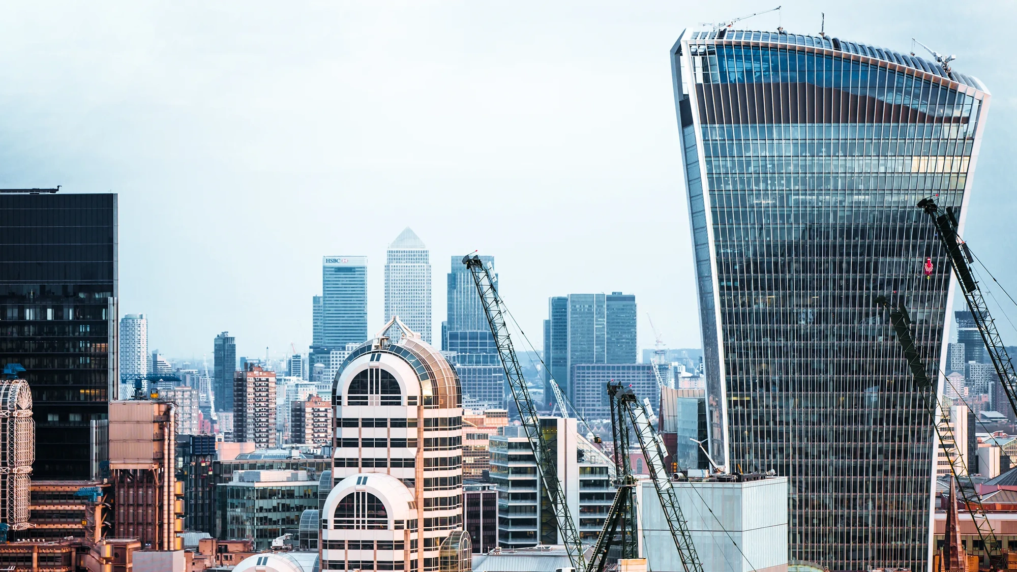 A view of the London skyline at dusk, bathed in a pinkish-golden light, with a cloudy sky. Four construction cranes are visible, hinting at ongoing construction work in the area.