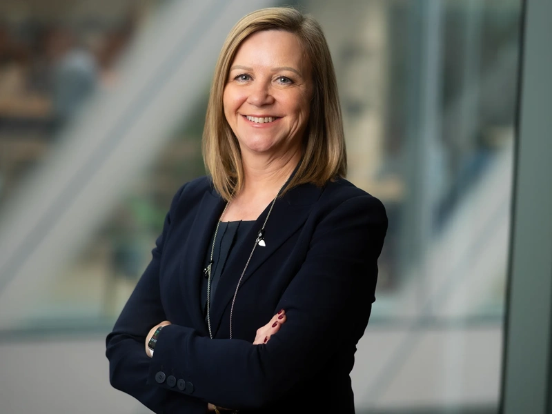 Patricia Moore, Managing Director, UK, standing in office space in front of a glass window. There are hints of greenery, suggesting some trees or plants.