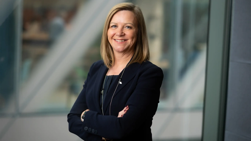 Patricia Moore, Managing Director, UK, standing in office space in front of a glass window. There are hints of greenery, suggesting some trees or plants.
