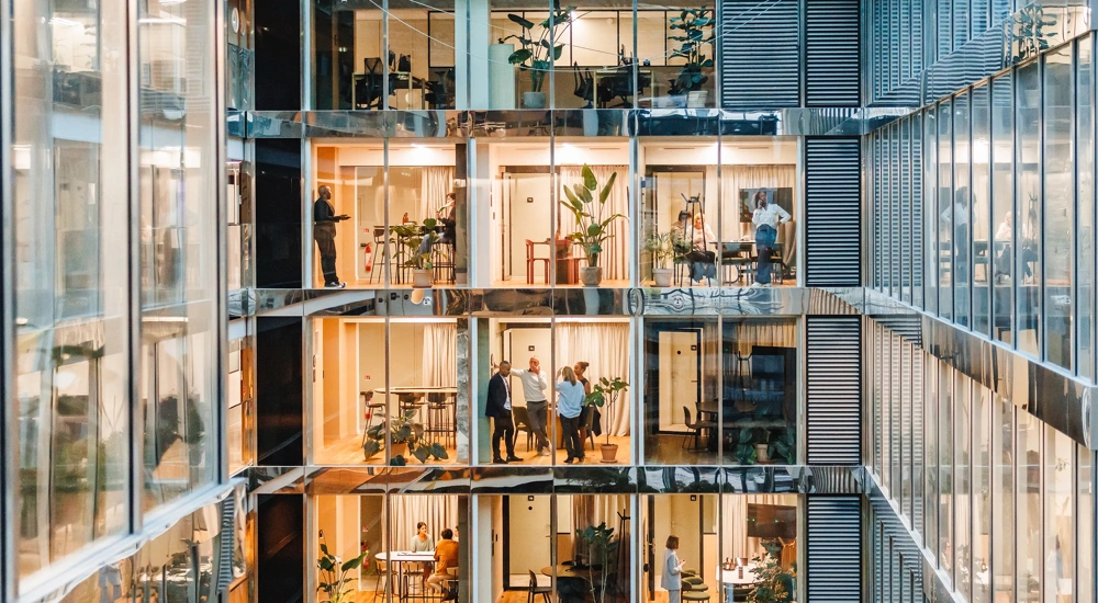 Men and women of various backgrounds collaborate and work independently in a spacious, well-lit office setting, each attired suitably for a professional setting, using modern tools and technology.