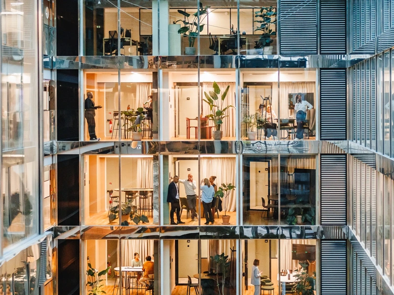Men and women of various backgrounds collaborate and work independently in a spacious, well-lit office setting, each attired suitably for a professional setting, using modern tools and technology.