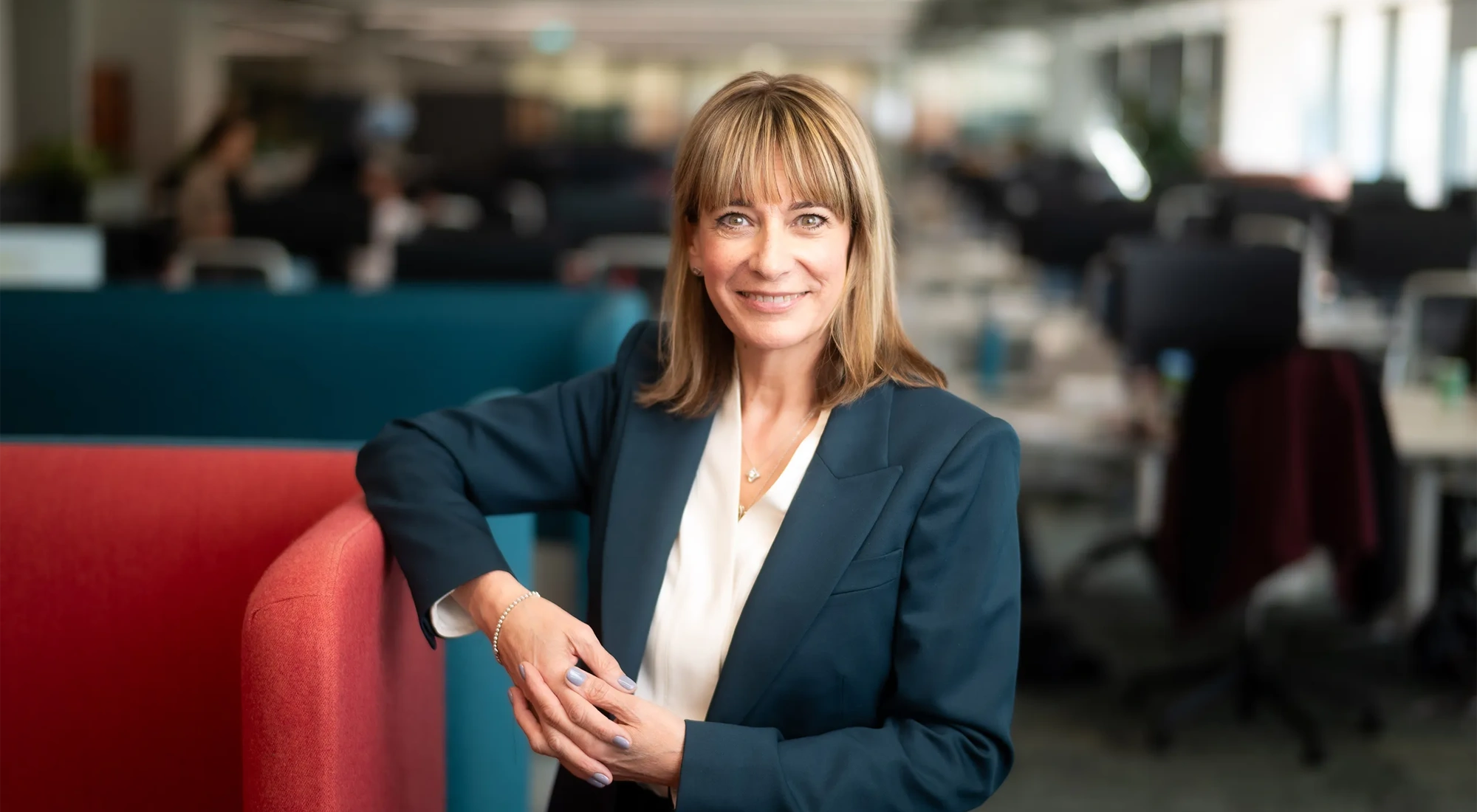Portrait of Emma Gilthorpe, Chief Transformation Officer, standing in office against red chair in blue suit.