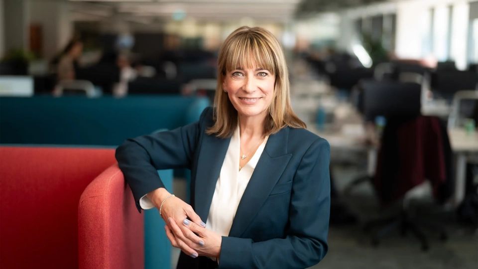 Portrait of Emma Gilthorpe, Chief Transformation Officer, standing in office against red chair in blue suit.