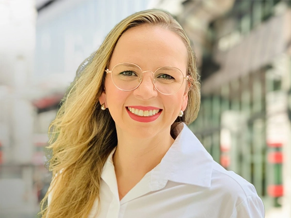 Portrait of Kamila Lima, Associate Director,standing outside in front of buildings featuring an urban cityscape with a mix of architectural styles. The scene includes some windows and red features, adding a touch of color.