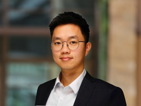 Issac Hou standing inside in front of a window displaying an outdoor urban setting, featuring modern architectural elements with clean lines. There are hints of greenery, suggesting some trees or plants.