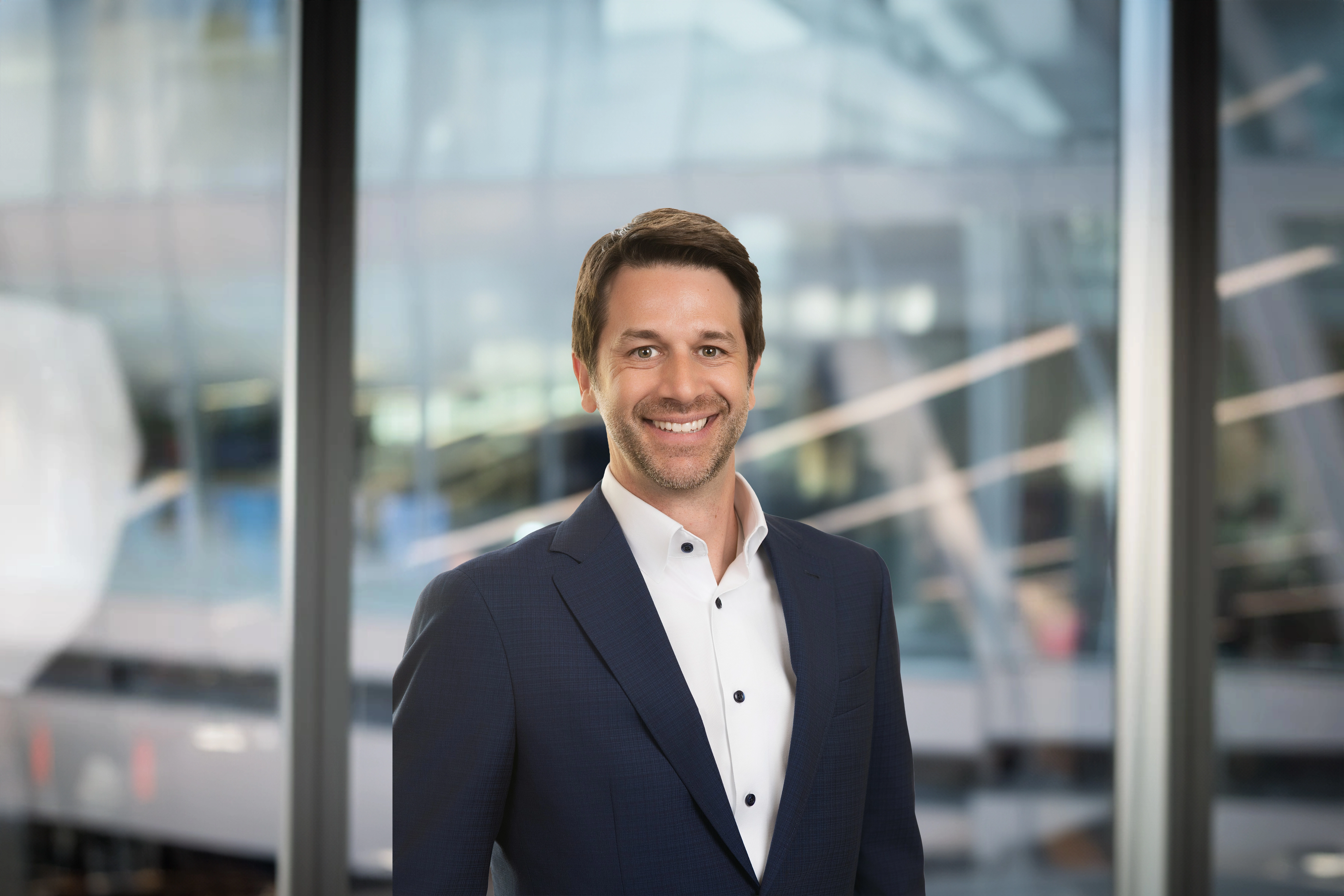 Portrait of Christopher Fields,  standing infront of windows in office with faint city buildings in the background smiling.