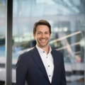 Portrait of Christopher Fields, standing infront of windows in office with faint city buildings in the background smiling.