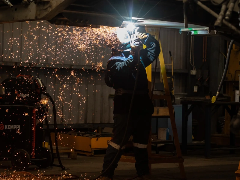 A welder in protective gear, including a welding helmet and gloves, is working in a dimly lit workshop. Bright orange sparks fly outward as they weld metal overhead, illuminating the area around them. Industrial equipment and tools are visible in the background, creating a rugged and workshop-like atmosphere