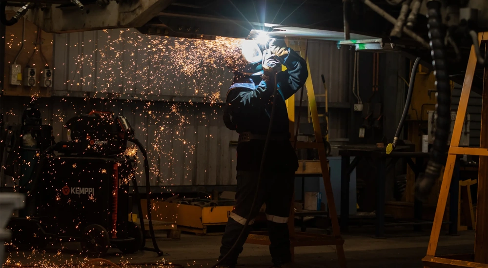 Welder in protective equipment works on a raised project in a dimly lit workshop, Australia