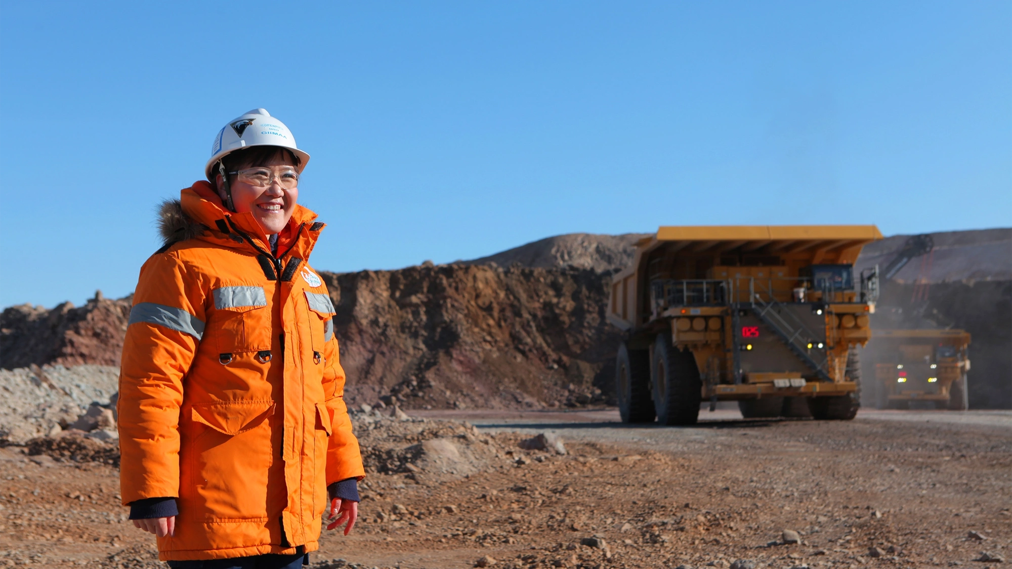Woman standing in sandy construction site wearing orange hi vis and helmet