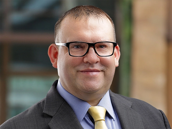 Portrait of Marc Fooks, UAE Infrastructure Lead, outside in front of a blurred brown background featuring a building and mix of natural and urban elements.