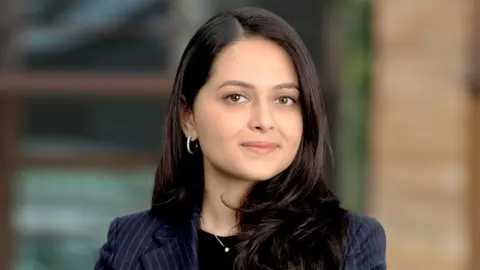 Portrait of Aparna Servansingh, Associate Director, outside in front of a blurred brown background featuring a building and mix of natural and urban elements.