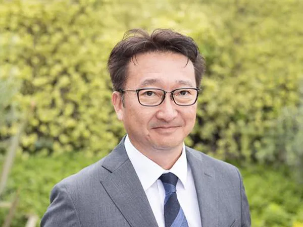 Portrait of Toshio Kosuge, Director, Clean Energy and Transmission, Japan, standing outside against green bushes, in grey suit smiling.