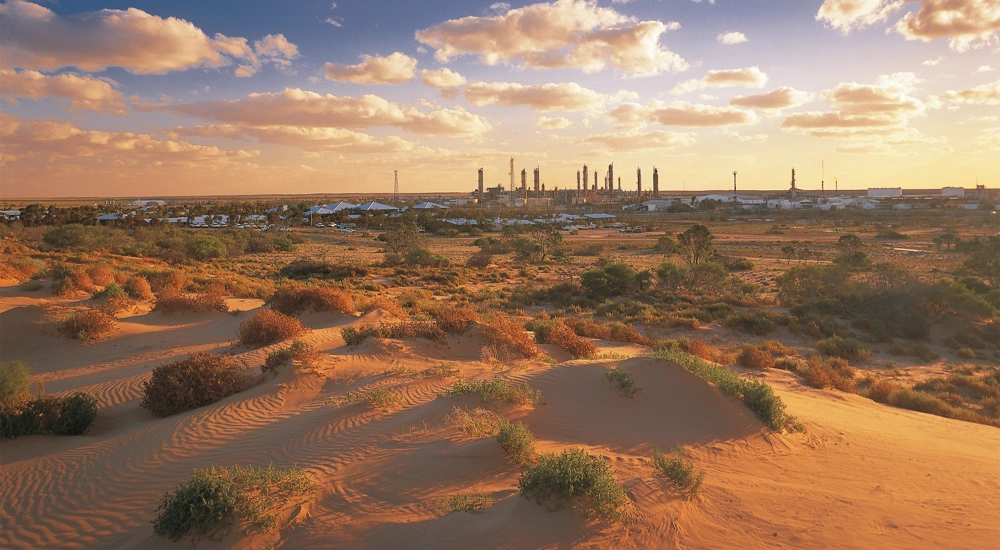 A sandy desert with some tufts of plants in the foreground of the image, with a clean energy plant far away in the distance.