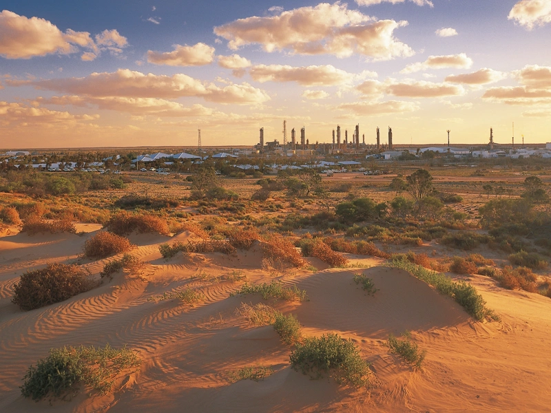 A sandy desert with some tufts of plants in the foreground of the image, with a clean energy plant far away in the distance.