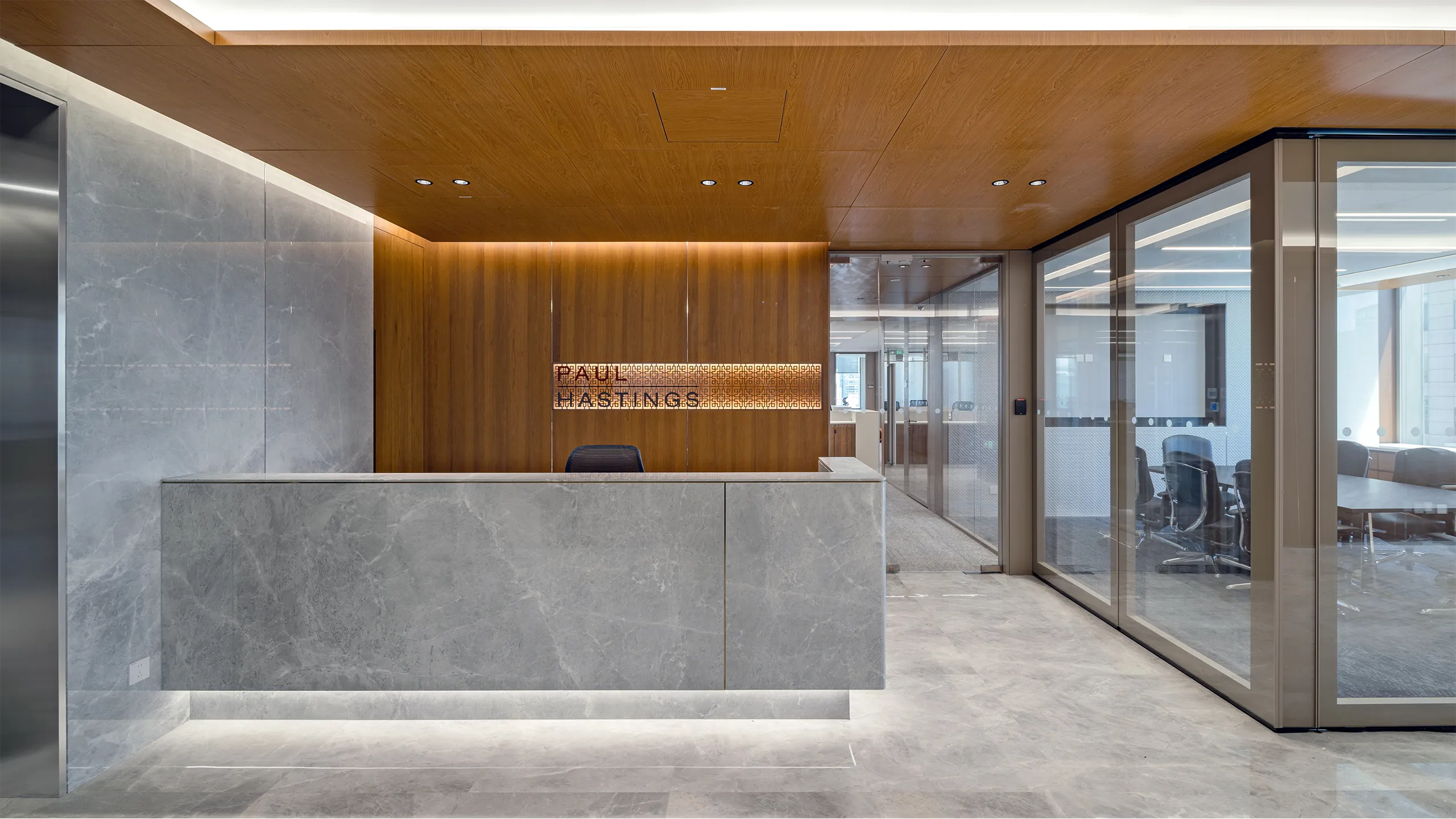 Modern office reception with a large stone desk, wood-panelled walls and ceiling, a backlit Paul Hastings sign, and glass-walled meeting rooms.