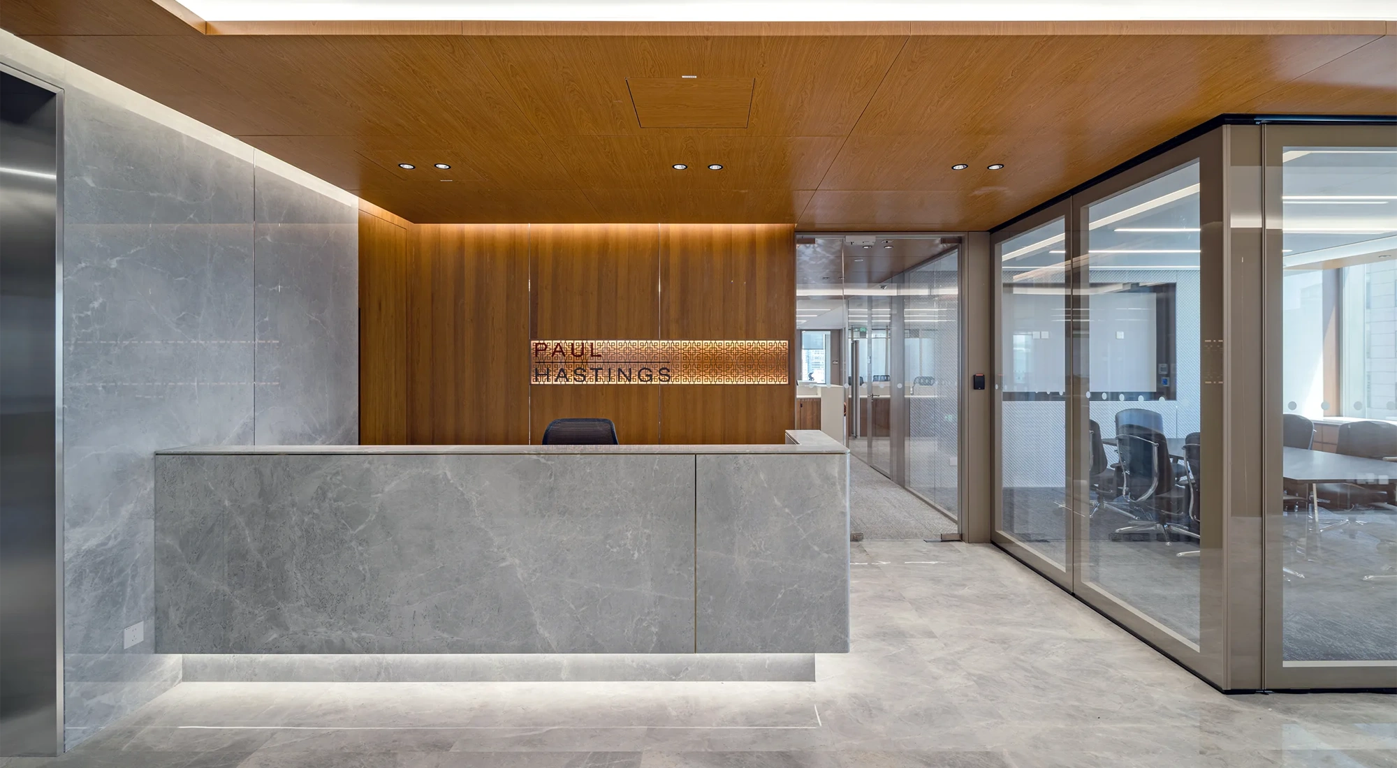 Modern office reception with a large stone desk, wood-panelled walls and ceiling, a backlit Paul Hastings sign, and glass-walled meeting rooms.
