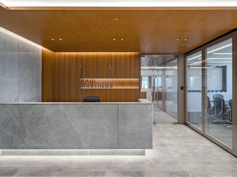 Modern office reception with a large stone desk, wood-panelled walls and ceiling, a backlit Paul Hastings sign, and glass-walled meeting rooms.