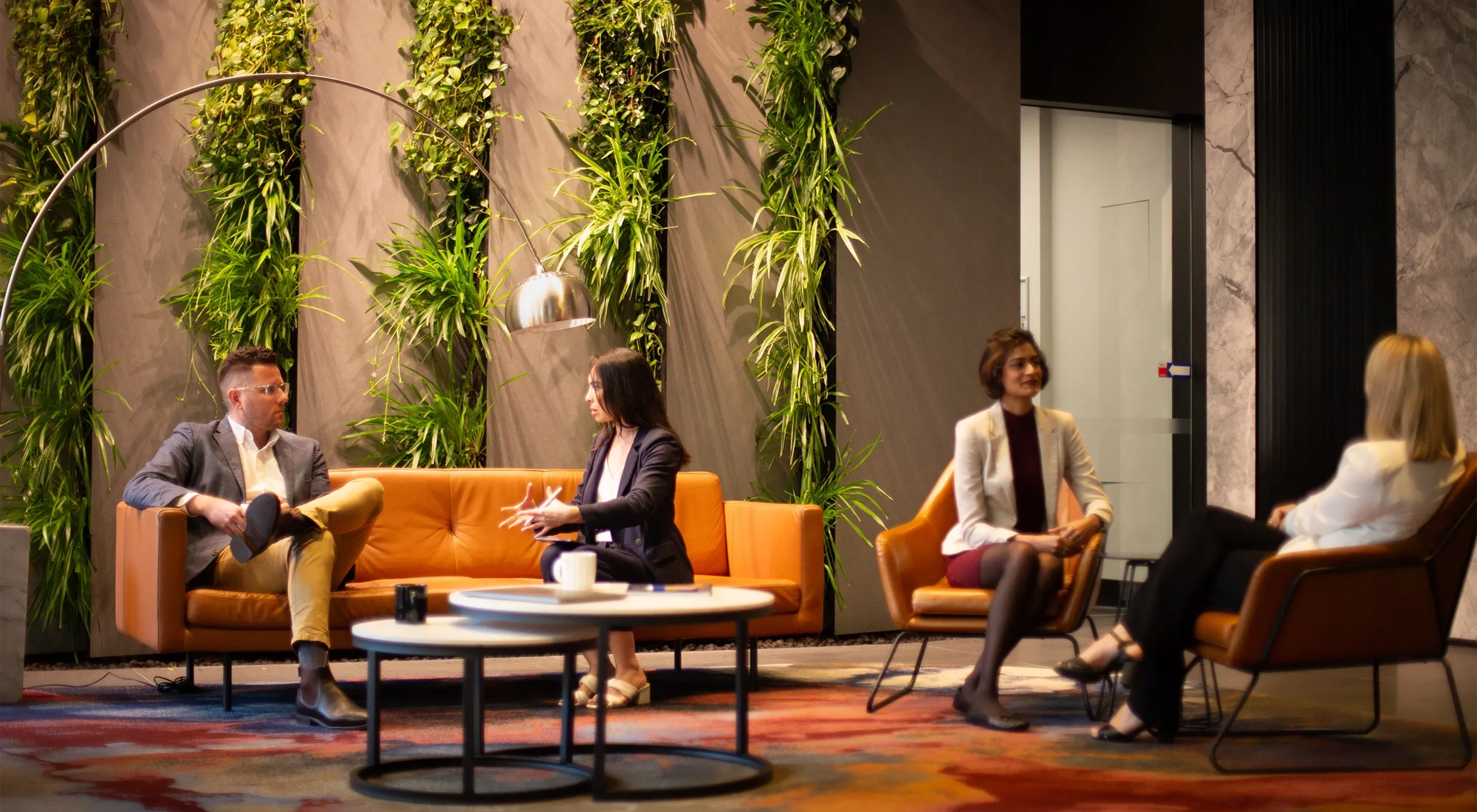 Group of four colleagues sitting separately in pairs on orange sofas in a plant-filled office.