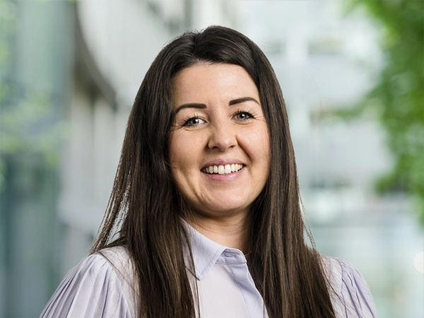 Portrait of Charlotte Treadwell, UK Communications Manager, standing outside in front of an outdoor urban setting, featuring modern architectural elements with clean lines. There are hints of greenery, suggesting some trees or plants.