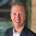 Josh Wentzell, outside in front of a blurred brown background featuring a building and mix of natural and urban elements.
