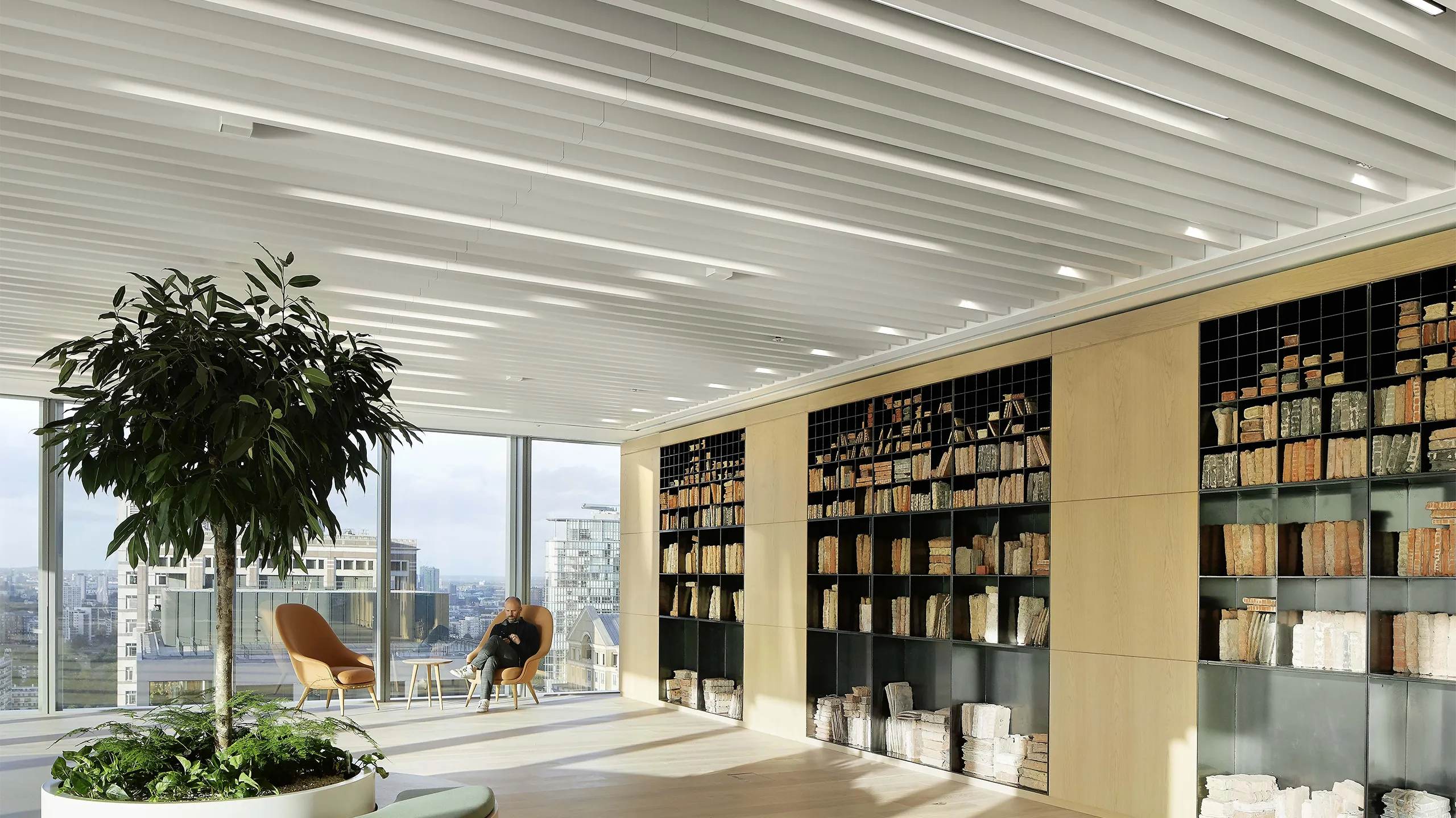 Bright plant filled office space with glass windows and shelf full of books. There's a man in the corner on a orange chair.