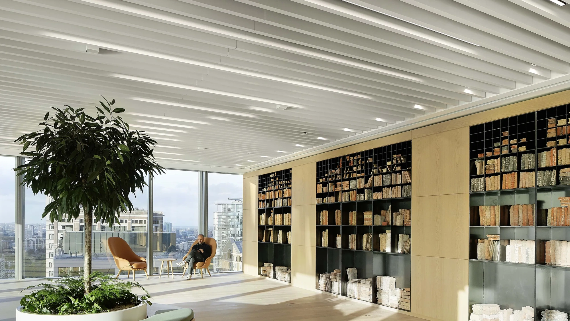 Bright plant filled office space with glass windows and shelf full of books. There's a man in the corner on a orange chair.